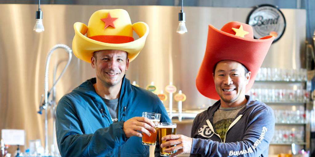 Manny and Roger wearing large novelty foam cowboy hats and toasting with pints of beer